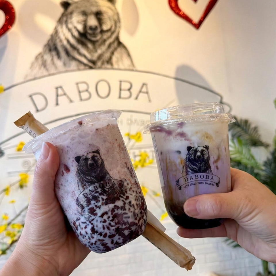 Two Daboba bubble tea drinks are held in front of a bear mural, surrounded by yellow blossoms and greenery.