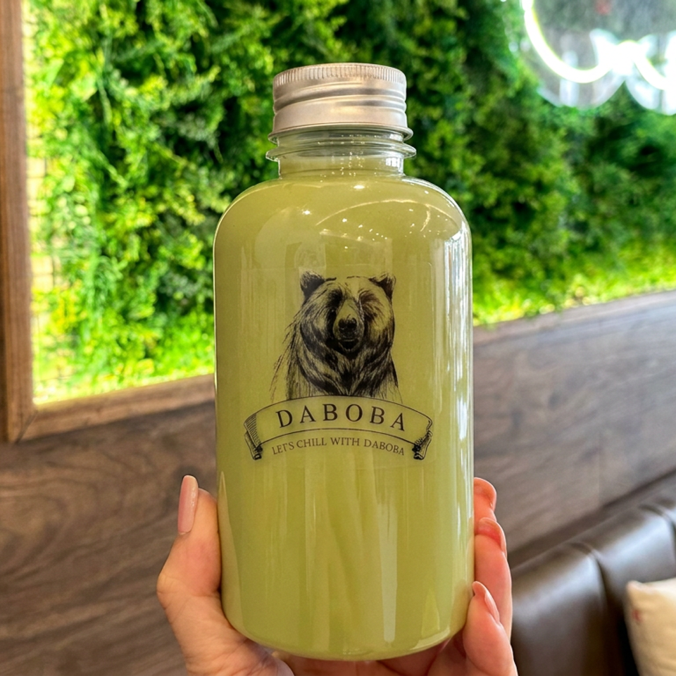 Hand holding a bottle of pale green Daboba drink in a cozy restaurant booth with greenery and wood paneling.