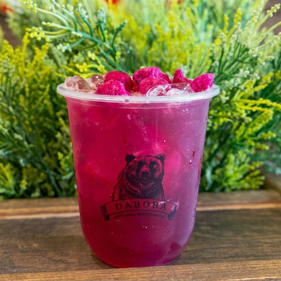 Bright magenta iced drink topped with raspberries in a branded Daboba cup, displayed against lush greenery on a wooden tray.