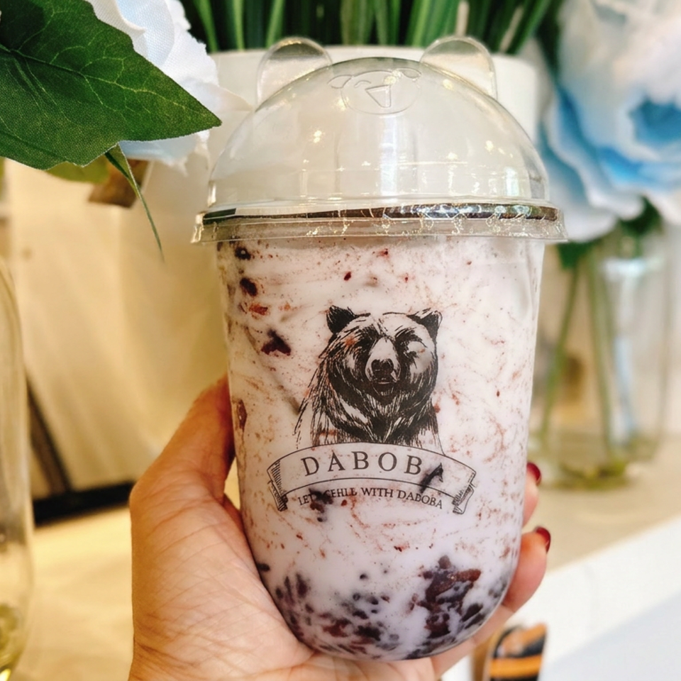 Hand holding a berry-speckled milk tea in a domed cup labeled DABOB, surrounded by blue and white flowers.