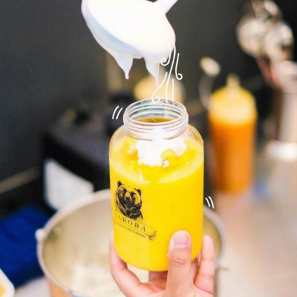 Hand holding a bright yellow fruit drink in a branded jar as whipped cream is added at a drink preparation counter.