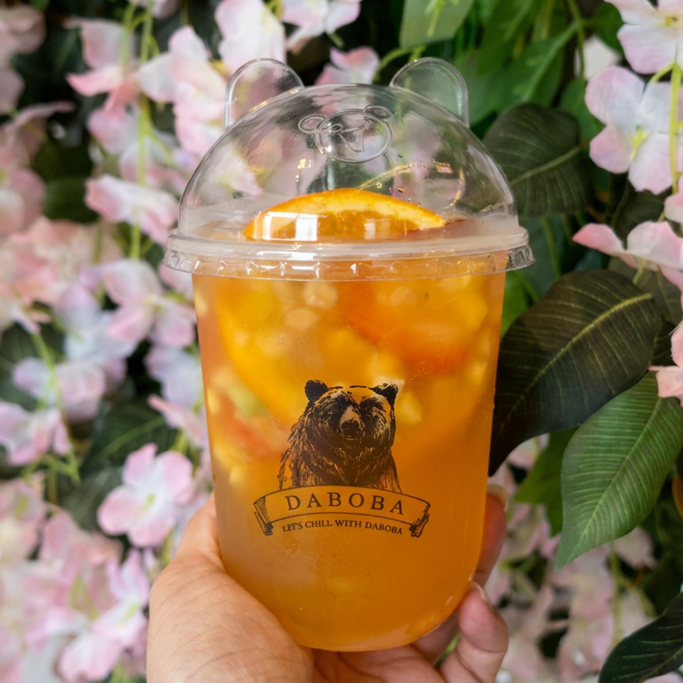 Hand holding a clear cup of orange fruit tea with citrus slices, branded Daboba logo, against a backdrop of pink flowers.