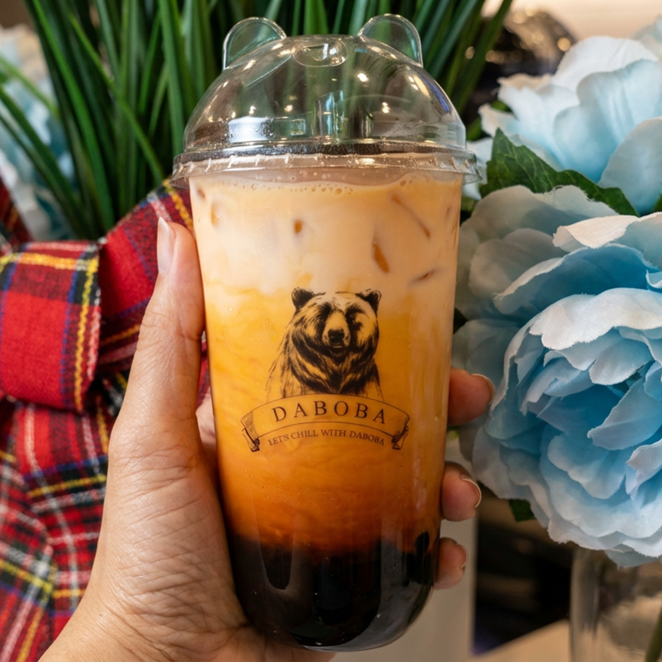 Hand holding a DABOBA iced milk tea with tapioca pearls, surrounded by blue flowers and a red plaid bow.